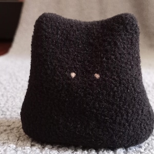 Könnte beinhalten: Ein schwarzes, katzenförmiges Stofftier mit zwei kleinen, hellfarbigen, gestickten Augen. Das Spielzeug hat ein abgerundetes, strukturiertes Aussehen und ein schlichtes Design. Das Katzenspielzeug liegt auf einer grau strukturierten Oberfläche.