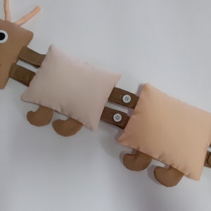 Pode incluir: Um brinquedo de lagarta marrom e bege com um rosto sorridente e dois olhos grandes. A lagarta é feita de tecido e tem uma série de segmentos conectados. Cada segmento tem um par de pernas marrons e um fecho de botão.