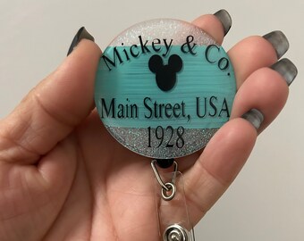 May include: A turquoise and silver glitter badge holder with a black Mickey Mouse silhouette and the text "Mickey & Co. Main Street, USA 1928".
