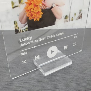 Peut inclure: Un support de lecteur de musique en acrylique transparent avec une photo d'une personne tenant un bouquet de fleurs jaunes. Le support a un bouton de lecture blanc, un bouton de rembobinage blanc et un bouton d'avance rapide blanc. Le texte "Lucky Jason Mraz (feat. Colbie Caillat)" est imprimé sur le support. L'heure "0:35" est affichée sur le côté gauche du support, et l'heure "3:11" est affichée sur le côté droit du support.