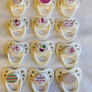May include: Twelve white pacifier clips with different colorful designs, including flamingos, fruit, and hearts. The clips are made of plastic and have a loop on the top for attaching to a pacifier.