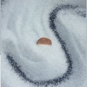 May include: A close-up shot of a pile of clear, sparkling sand with a copper penny partially buried in it. The sand is arranged in a swirling pattern against a dark background. The penny is approximately 0.75 inches in diameter.