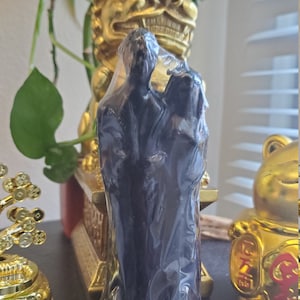 Pode incluir: Uma vela preta com duas figuras de pé lado a lado. As figuras estão em uma pose de abraço. A vela está sobre uma mesa com uma estátua dourada e uma figura de sapo dourada.