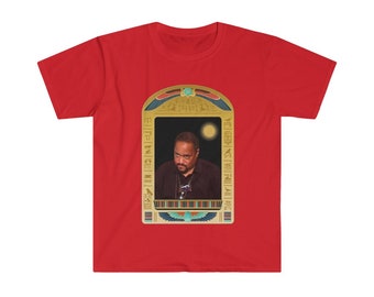 Puede incluir: Camiseta roja con una imagen enmarcada en oro de un hombre con barba que lleva una camisa negra. La imagen está rodeada de jeroglíficos y un sol.