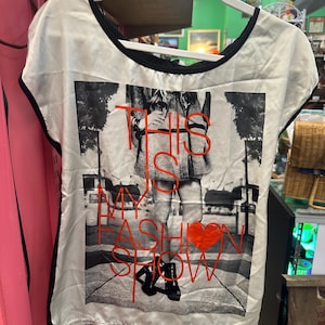 May include: Cream-coloured short-sleeved top with black trim and a graphic print. The print shows a monochrome image of a person with the text "THIS IS MY FASHION SHOW" in red, plus a red heart. The top is on a white hanger.