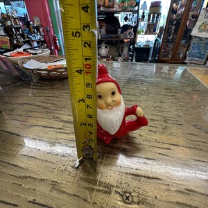 May include: A small, red and white ceramic gnome figurine. The gnome has a white beard, a red hat, and is approximately 3 inches tall. The figurine is on a wooden surface.