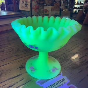 May include: A bright green glass pedestal bowl with a frilled edge and floral patterns. The bowl is lit, highlighting its distinctive colour and design. The sticker reads "MOTIVATION determines what you do ATTITUDE determines how well you do it."