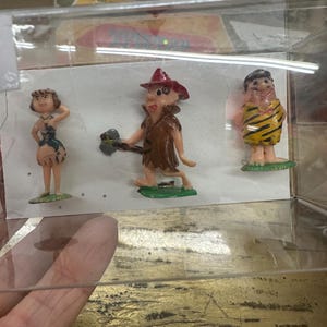 May include: A set of three vintage Flintstones figurines displayed in a clear plastic case. The figurines depict Wilma, Fred, and Barney in their classic cartoon attire. The figures are painted with bright colours and stand on small green bases.