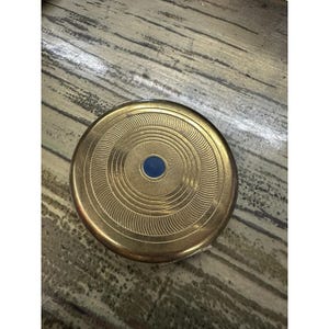 May include: A round gold-toned metal compact case featuring a textured surface with concentric circular patterns and a small, solid dark blue circular inlay centered on the lid. The object has a polished metallic finish and a circular shape.