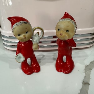 Könnte beinhalten: Zwei rote und weiße Porzellan-Weihnachtswichtelfiguren. Ein Wichtel hält eine goldene und weiße Trompete, der andere Wichtel hält sich die Hand vor den Mund.