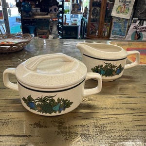 May include: A vintage cream-colored sugar bowl with a lid and a creamer. Both pieces have a black trim and a floral design with blue and green accents. The sugar bowl has a handle on the lid and two side handles.