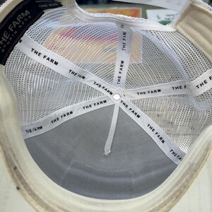 May include: Inside view of a white trucker hat with a mesh back and a curved brim. The hat features the text "THE FARM" on the interior bands and a label that reads "GOORIN BROS." and "ONE SIZE".