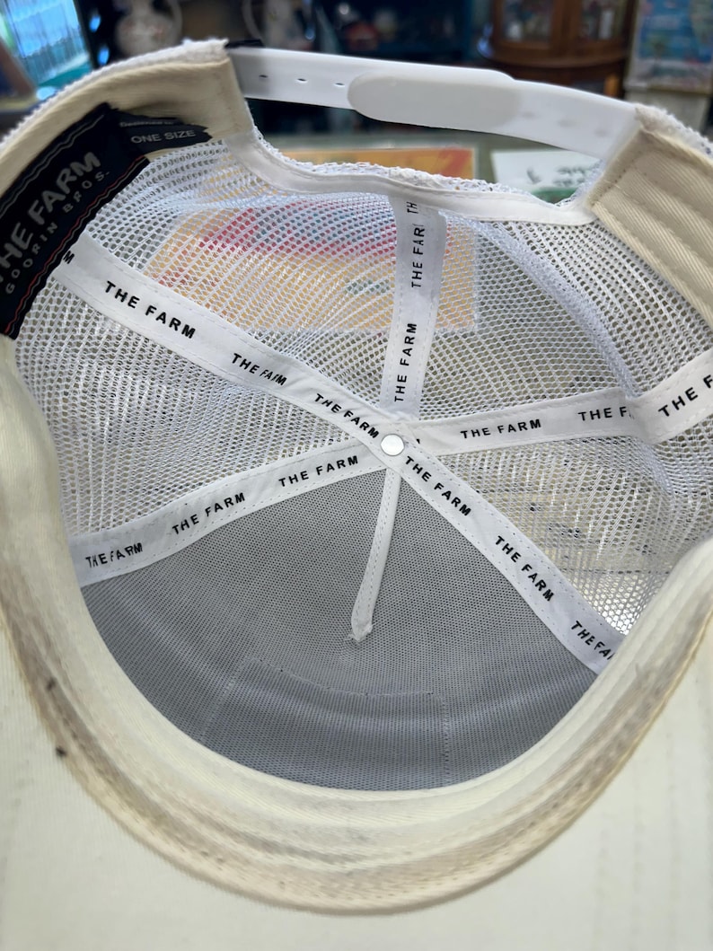 May include: Inside view of a white trucker hat with a mesh back and a curved brim. The hat features the text "THE FARM" on the interior bands and a label that reads "GOORIN BROS." and "ONE SIZE".