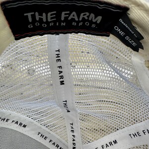 May include: Cream-colored baseball cap with a white mesh back and a black label that reads "THE FARM GOORIN BROS." The interior features white straps with "THE FARM" printed repeatedly. The tag also says "ONE SIZE."