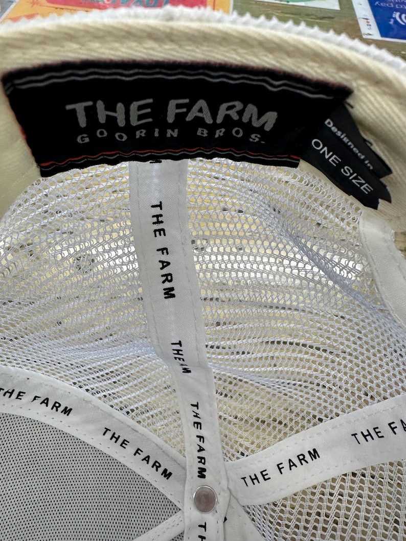 May include: Cream-colored baseball cap with a white mesh back and a black label that reads "THE FARM GOORIN BROS." The interior features white straps with "THE FARM" printed repeatedly. The tag also says "ONE SIZE."