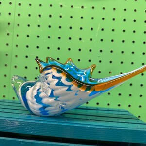 May include: A decorative glass seashell sculpture with a blue, white, and orange color scheme. The sculpture has a detailed, spiky design and sits on a teal wooden surface. The background is a green pegboard.