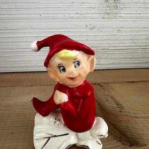 May include: A vintage ceramic elf figurine. The elf wears a bright red outfit and hat with a white pom-pom. The figurine has a smiling face, blue eyes, and blonde hair. It sits on a white base with gold trim, perfect for holiday decor.