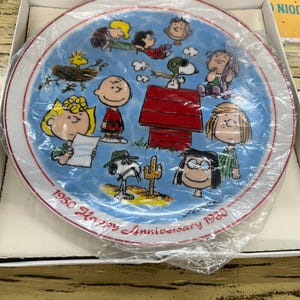May include: A ceramic plate featuring a colorful illustration of Peanuts characters. The plate has a blue background with a red border and the text "1950 Happy Anniversary 1980". The plate is in its original packaging.