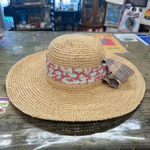 以下が含まれることがあります： 幅広のつばが付いた、織り込まれた麦わら帽子。 ピンクと白のサンゴ模様のバンドと、チェック柄のリボンが特徴です。 日焼け防止用に設計されており、スタイリッシュなアクセサリーです。