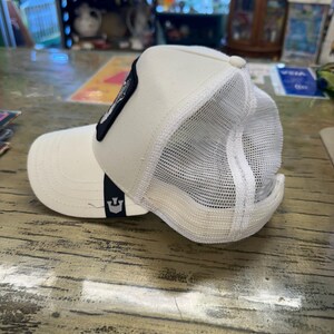 May include: Cream-colored trucker hat with a white mesh back and a curved brim. A black patch with a design is on the front. The hat has a snap closure and a small logo on the side.