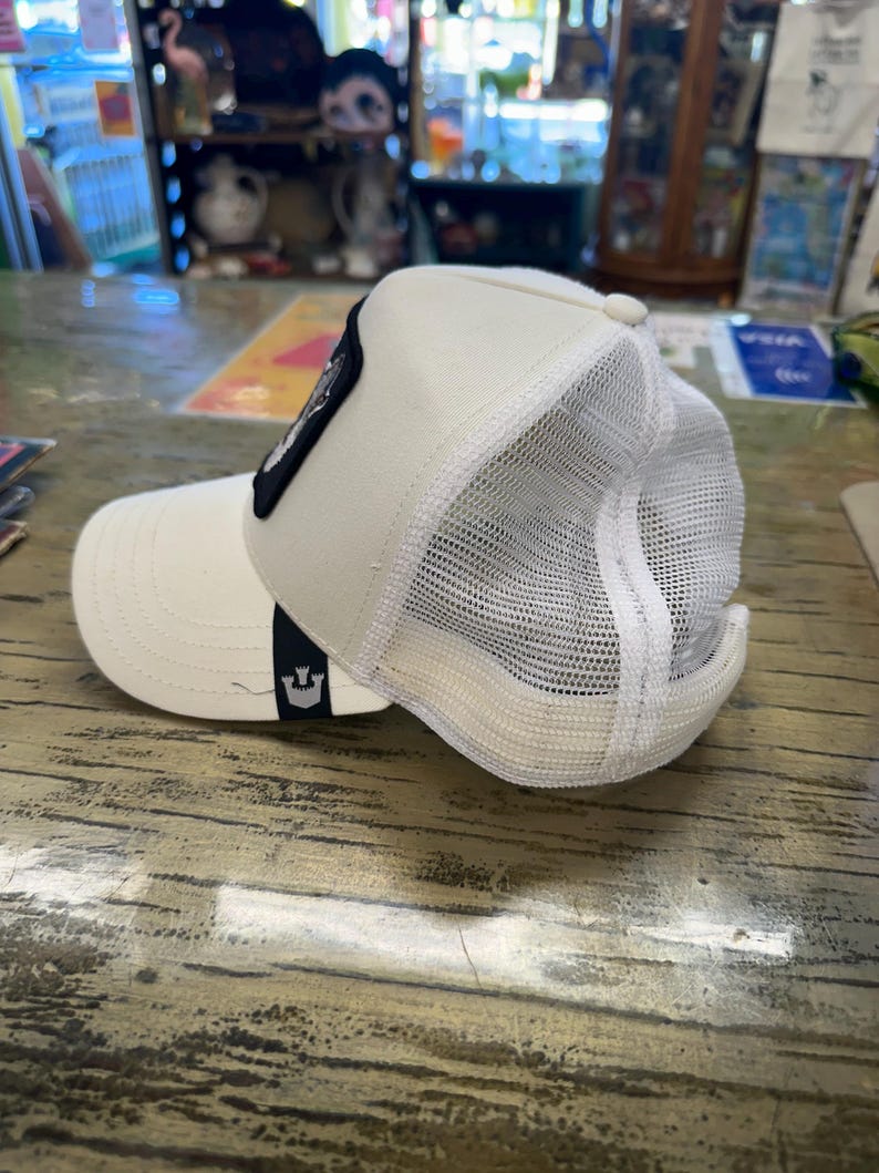 May include: Cream-colored trucker hat with a white mesh back and a curved brim. A black patch with a design is on the front. The hat has a snap closure and a small logo on the side.