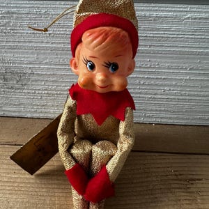 Puede incluir: Un adorno de elfo navideño dorado y rojo con una campana en su gorro. El elfo está sentado con las piernas cruzadas y tiene una expresión amistosa en su rostro.