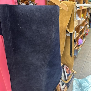 May include: A navy blue suede garment hangs on a wooden hanger, next to a tan suede garment with a zipper. The garments are displayed in a vintage shop setting, with shelves and other items visible in the background.