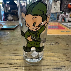 May include: A clear glass tumbler featuring a cartoon image of Elmer Fudd in a green hunting outfit. The character is holding a rifle and wearing a green hat. The words "Elmer Fudd" are printed at the bottom of the glass.