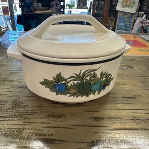 May include: Cream-colored ceramic casserole dish with a lid and handle. The dish features a black band and a decorative border of green leaves, blue and green fruits. The dish is sitting on a wooden surface.