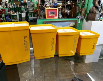 May include: A set of four yellow vintage kitchen canisters with white lids. The canisters are labeled "FLOUR", "SUGAR", "COFFEE", and "TEA" in a vertical font. The canisters are arranged in a row on a glass surface.