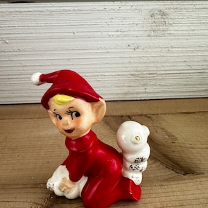 May include: A red and white ceramic elf figurine sitting with a white ceramic bird on its back. The elf is wearing a red hat with a white pom-pom.