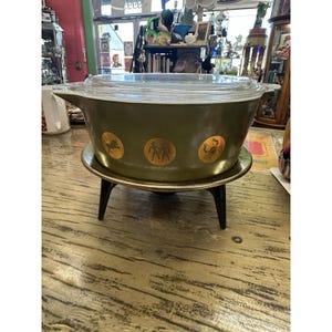 May include: An olive green glass casserole dish with a clear glass lid, featuring a series of gold circular zodiac symbols around the exterior. The dish is supported by a dark brown metal stand with three tapered legs and a circular base.