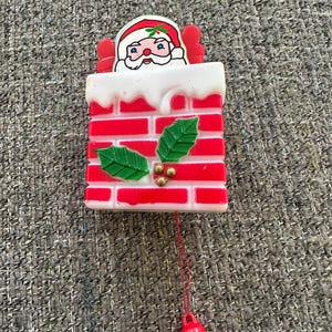 May include: A plastic Christmas ornament featuring a Santa Claus figure emerging from a red and white brick chimney. The chimney is decorated with green holly leaves and small gold berries. A red plastic bell hangs from a thin red string below.