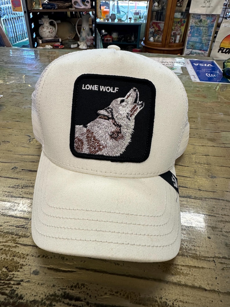 May include: A cream-colored baseball cap with a mesh back and a curved brim. The front features a black square patch with the words "LONE WOLF" above a howling wolf illustration. The cap is designed with a classic six-panel construction.
