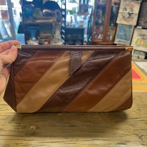 Puede incluir: Un bolso de mano vintage con un patrón de rayas diagonales en tonos marrones y beige. El bolso tiene forma rectangular con un cierre y ribete dorados. El bolso está hecho de cuero.