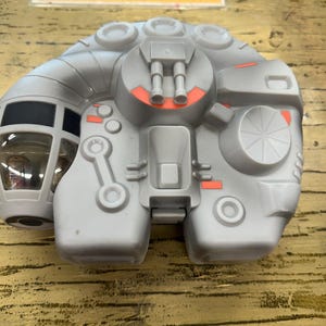 Può includere: Un'astronave giocattolo grigia con dettagli arancioni. Il giocattolo presenta un abitacolo, diversi elementi circolari e rettangolari e un design circolare sul lato. Il testo "JOIN OUR" è visibile sullo sfondo.