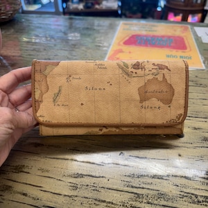 May include: A tan leather wallet with a world map design. The wallet is rectangular with a fold-over flap. The map features continents and islands in shades of brown and tan. The wallet is held by a person's hand.
