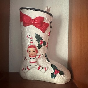 May include: A white ceramic Christmas stocking with a red bow, candy canes, holly, and an elf figurine. The stocking has a textured surface and a black rim. A festive holiday decoration.