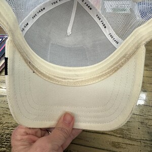 May include: Cream-colored baseball cap with a white mesh back and a curved brim. The interior features a white band with the text "THE FARM" repeated. The cap is held by a hand, showcasing the underside of the hat.