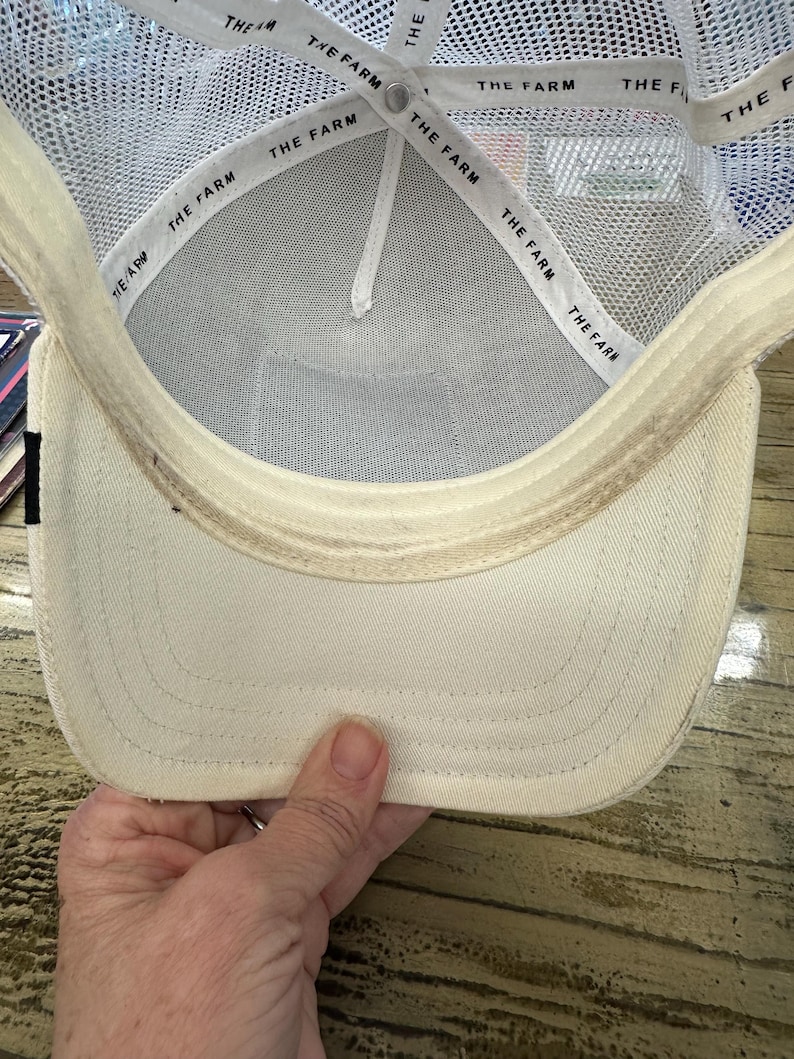 May include: Cream-colored baseball cap with a white mesh back and a curved brim. The interior features a white band with the text "THE FARM" repeated. The cap is held by a hand, showcasing the underside of the hat.