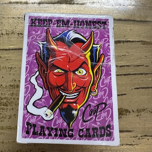Könnte beinhalten: Ein Kartenspiel mit einer Illustration eines roten Teufels, der eine Zigarre raucht. Der Text "KEEP 'EM HONEST" und "PLAYING CARDS" sind auf einem lila Hintergrund zu sehen.