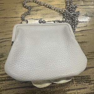 Peut inclure: Un petit sac &agrave; main en cuir blanc cass&eacute; avec une cha&icirc;ne argent&eacute;e. Le sac &agrave; main a une fermeture &agrave; fermoir et une surface textur&eacute;e. La cha&icirc;ne est faite de petits maillons m&eacute;talliques. Le sac est pos&eacute; sur une surface en bois.