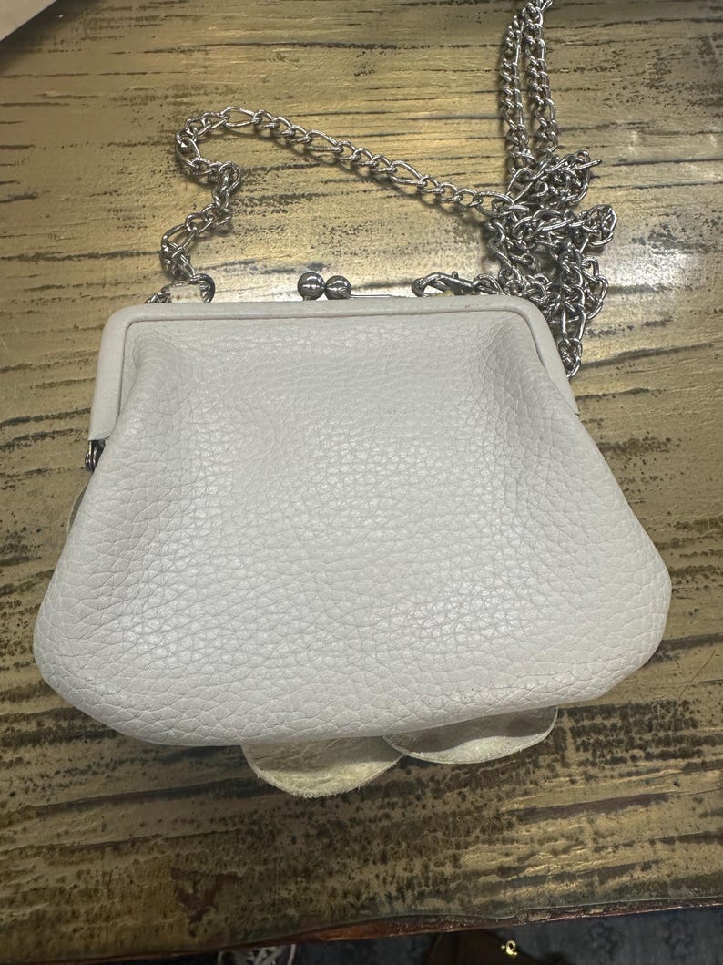 Peut inclure: Un petit sac &agrave; main en cuir blanc cass&eacute; avec une cha&icirc;ne argent&eacute;e. Le sac &agrave; main a une fermeture &agrave; fermoir et une surface textur&eacute;e. La cha&icirc;ne est faite de petits maillons m&eacute;talliques. Le sac est pos&eacute; sur une surface en bois.