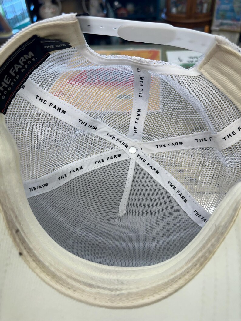 May include: A white trucker hat with a mesh back and a curved brim. The interior features white bands printed with "THE FARM". The hat has a white adjustable strap and a tag that reads "ONE SIZE".