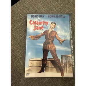 May include: A DVD case for the film Calamity Jane starring Doris Day and Howard Keel. The cover features a person in a brown fringed outfit and hat standing between two wooden barrels against a blue sky. The text reads Calamity Jane.