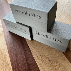 May include: Three grey rectangular boxes stacked on a wooden surface. Each box is labelled "Needle Box" in embossed lettering. The boxes have a textured surface and a simple, modern design.