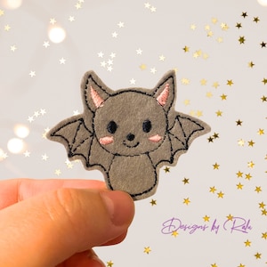 Könnte beinhalten: Eine graue Filzfledermaus mit rosa Wangen und schwarzen Augen. Die Fledermaus hat gestickte Flügel und einen niedlichen, cartoonartigen Ausdruck.