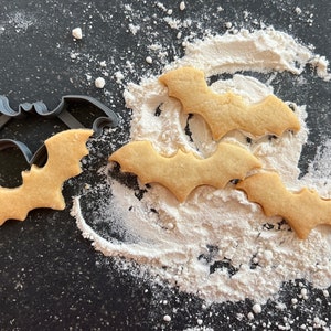 Puede incluir: Galletas con forma de murciélago y un cortador sobre una superficie oscura con harina. Las galletas son de color marrón claro y tienen un diseño detallado de alas de murciélago. El cortador es gris y tiene forma de murciélago.