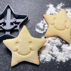 Puede incluir: Un cortador de galletas en forma de estrella gris junto a dos galletas en forma de estrella con caras sonrientes. Las galletas son de color marrón claro y el cortador es gris. La harina está esparcida alrededor de las galletas sobre una superficie oscura.