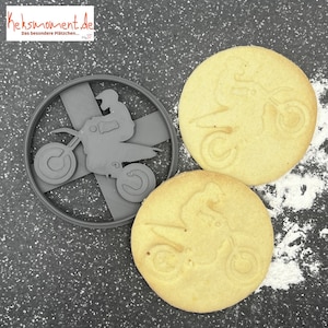 Peut inclure: Deux biscuits sablés avec un motif de pilote de motocross. Les biscuits sont de couleur brun clair et ont un motif en relief. Les biscuits sont sur une surface grise avec de la poudre blanche.
